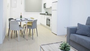 A modern kitchen with white cabinets, a dining area with a table and chairs, and a grey sofa. - IBERICABEDS CENTER 1 BEDROOM (Granada)