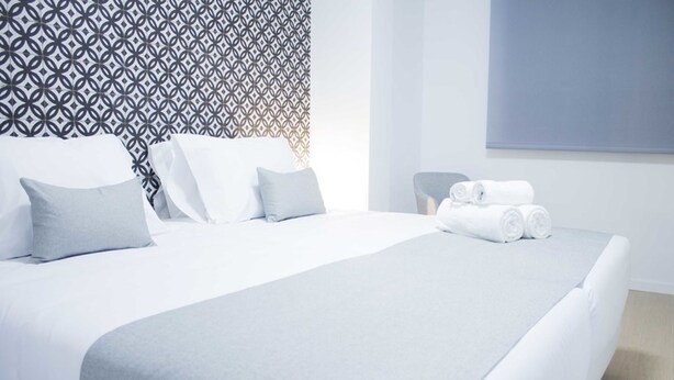 A neatly made bed with white linens and pillows, a chair, and rolled towels on a clean, modern room with a geometric patterned wall. - IBERICABEDS CENTER 1 BEDROOM (Granada)