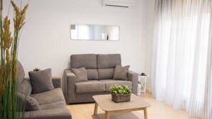 Een moderne woonkamer met een grijze bank, een houten salontafel met een plant, en een witte gordijn.