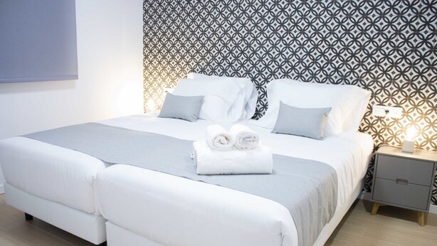 Een netjes opgemaakt bed met witte lakens en kussens, een nachtkastje met een lamp, en een behang met patroon achter het bed.