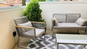Een balkon met twee rieten stoelen, een bank, een klein tafeltje en een vloerkleed met dessin.