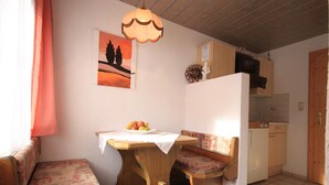 A small kitchen with a refrigerator, microwave, and wooden cabinets. There is a table with a white tablecloth and a fruit bowl. A bench with a floral cushion is also visible. - Haus Hrabovsky (Laengenfeld)