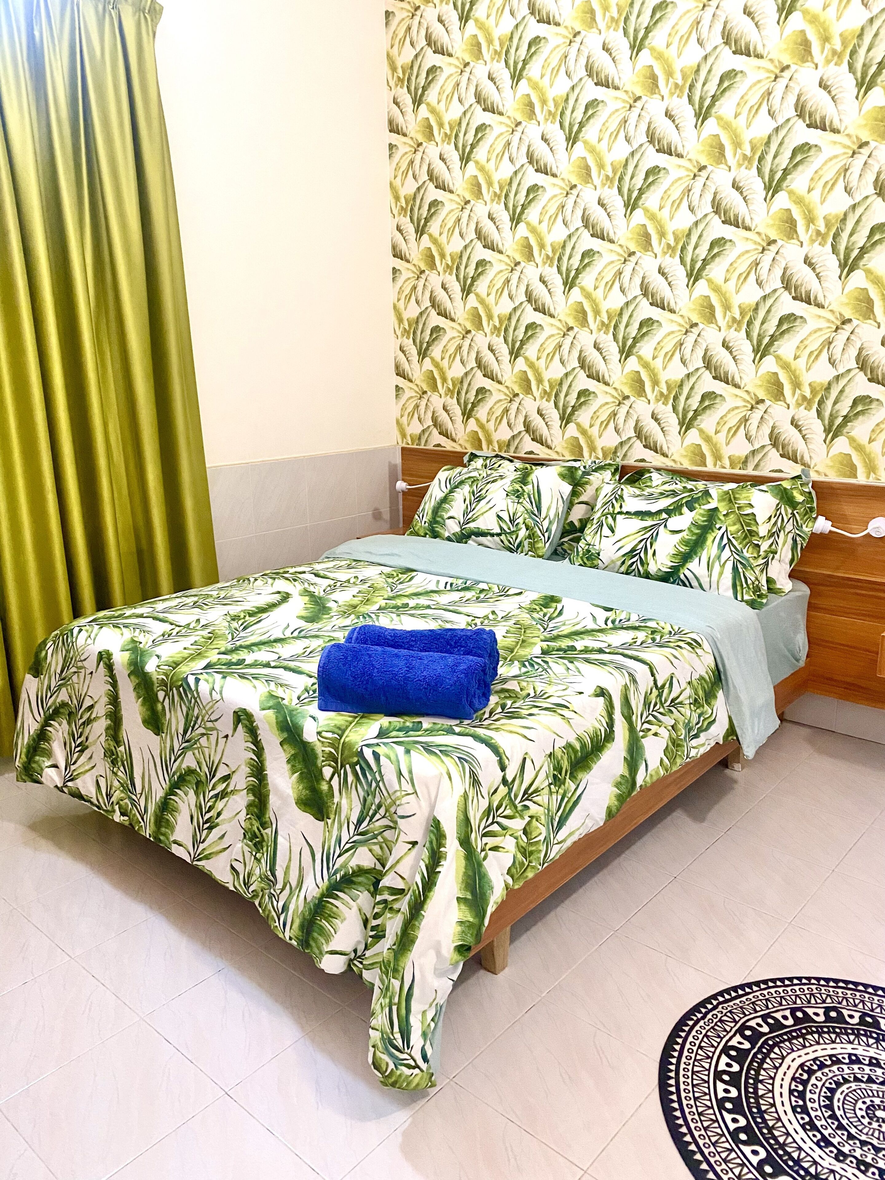 A bed with a tropical leaf pattern bedspread, a blue towel, and a patterned rug on the floor.