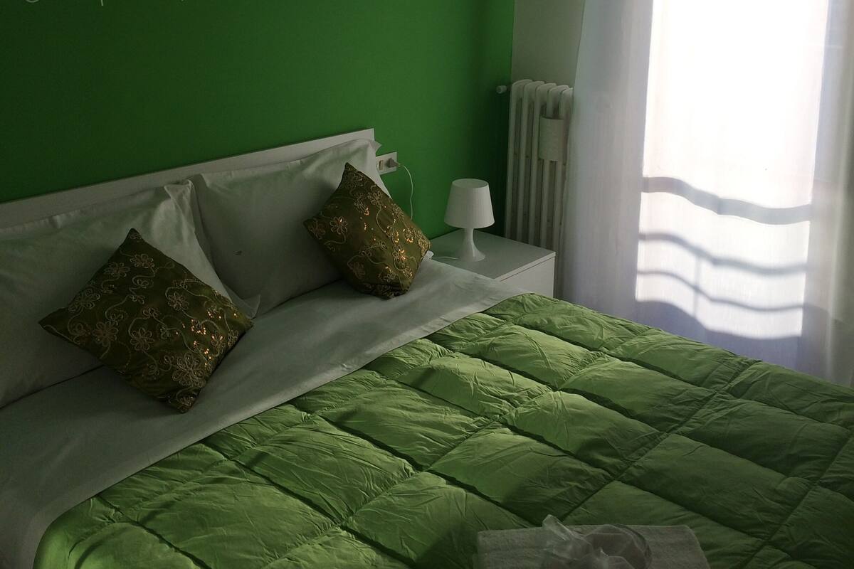 A neatly made bed with green bedding, a white bedside table with a lamp, and decorative pillows.