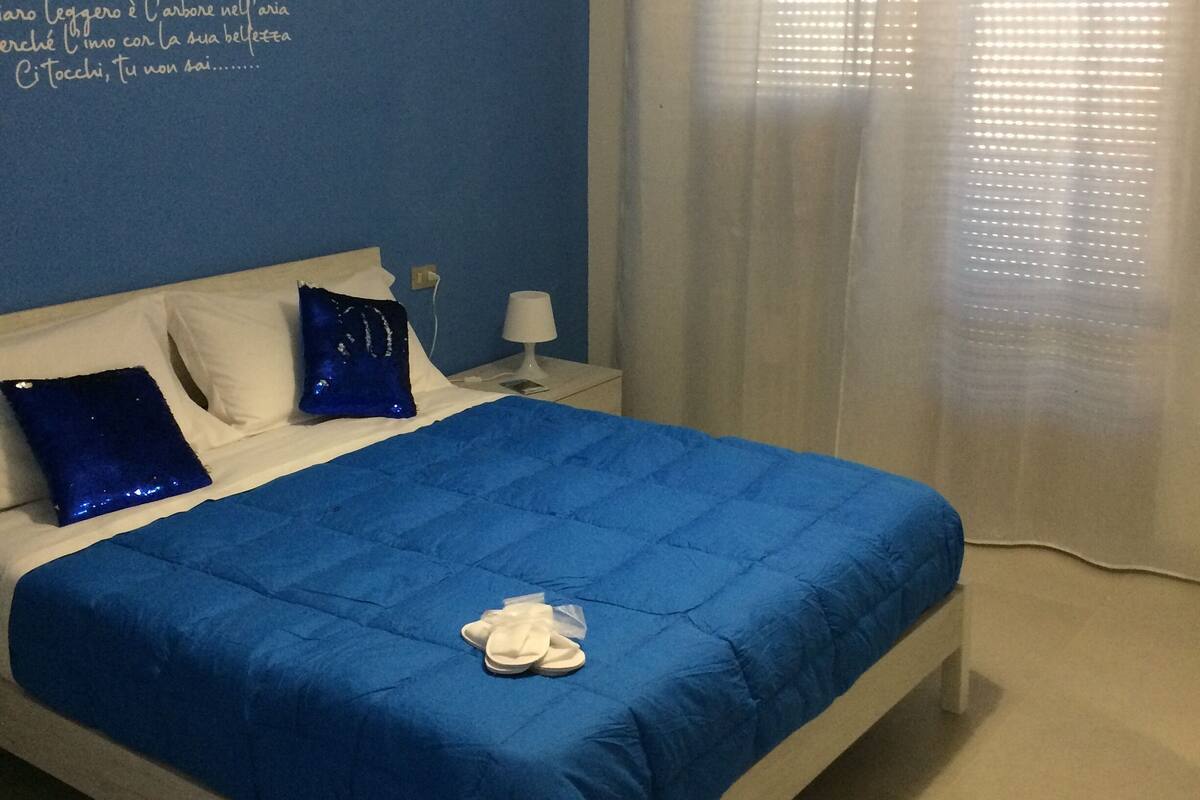A bedroom with a blue bedspread, white pillows, a white nightstand, a lamp, and a window with sheer curtains.