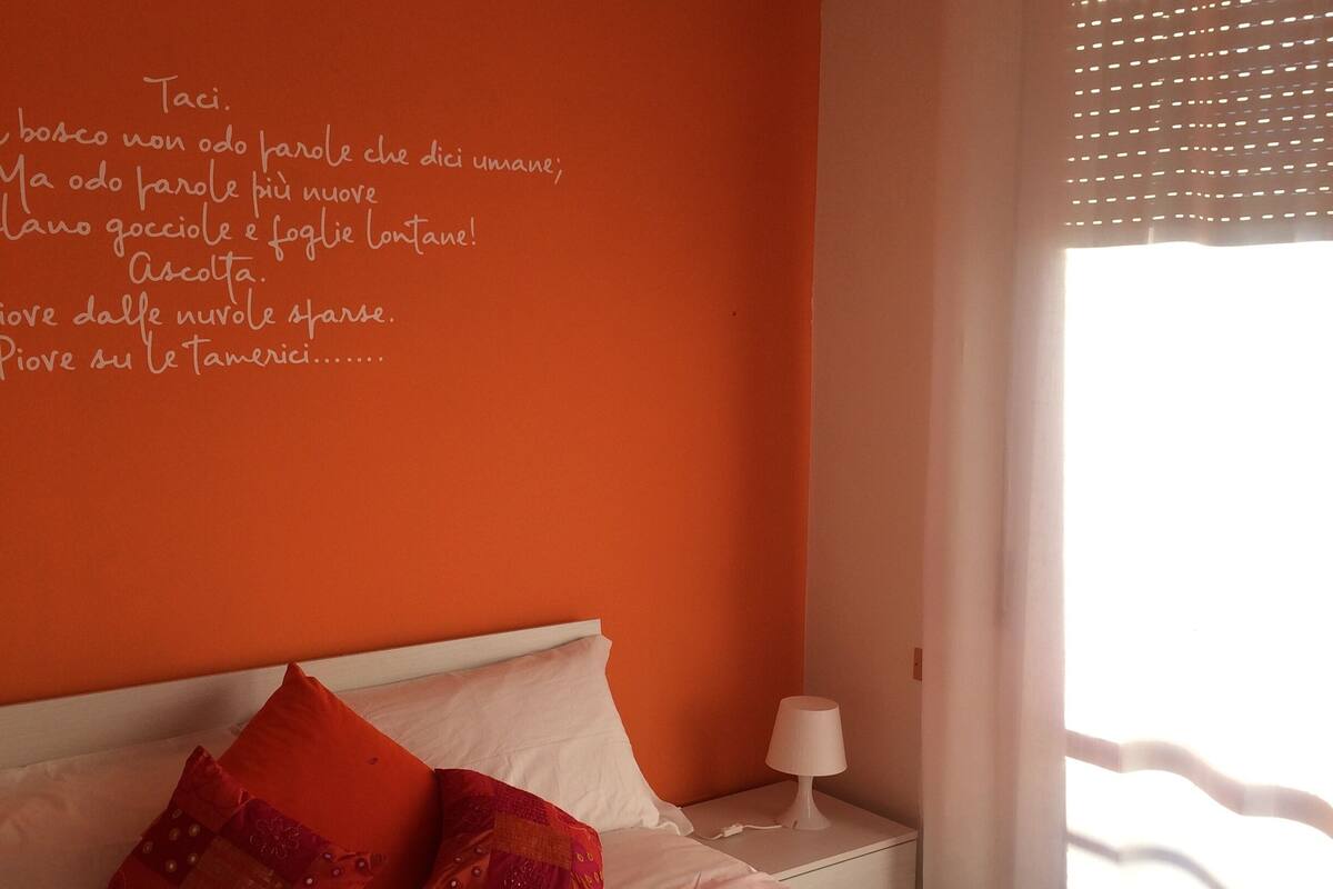 A bedroom with an orange wall, a white ceiling lamp, a bed with an orange quilt, and a window with a white curtain.