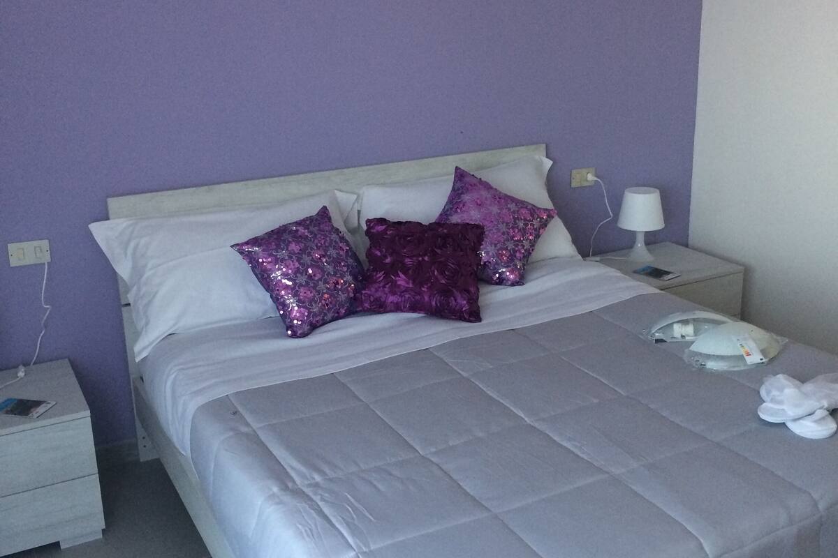 A neatly made bed with a grey bedspread and two purple pillows, a white nightstand with a lamp, and a wall with a motivational quote in Italian.