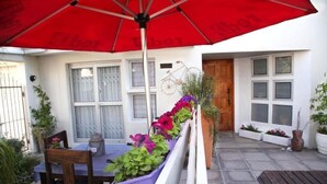 Un patio al aire libre con una sombrilla roja, una mesa de madera y sillas. Hay plantas en macetas y una bicicleta montada en la pared.