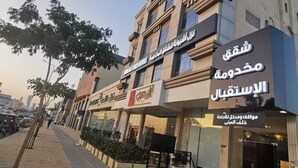A hotel with a sign in Arabic and English, a storefront with a sign in Arabic, and a paved sidewalk with trees and a parked motorcycle. - ashbona hotel suites (Riyadh)
