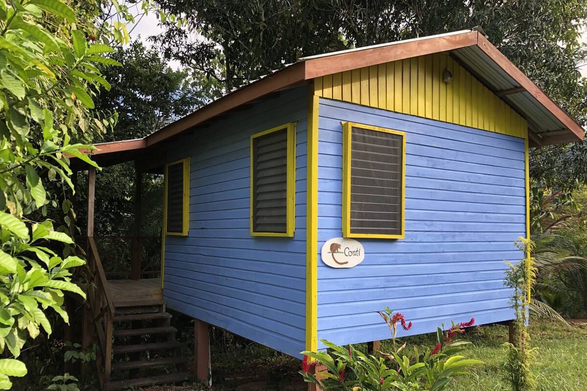 Un pequeño edificio de una sola planta con fachada azul y detalles amarillos, que cuenta con una escalera de madera que conduce a un porche cubierto.