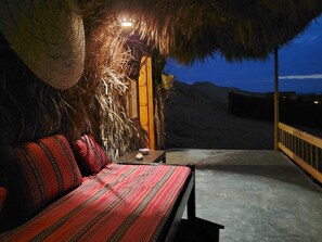 A bed with a red and black patterned bedspread, a nightstand with a bowl, and a thatched roof structure with a light source.