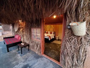 A room with a thatched ceiling, a bed with red and white bedding, a small table, and a woven basket hanging on the wall.