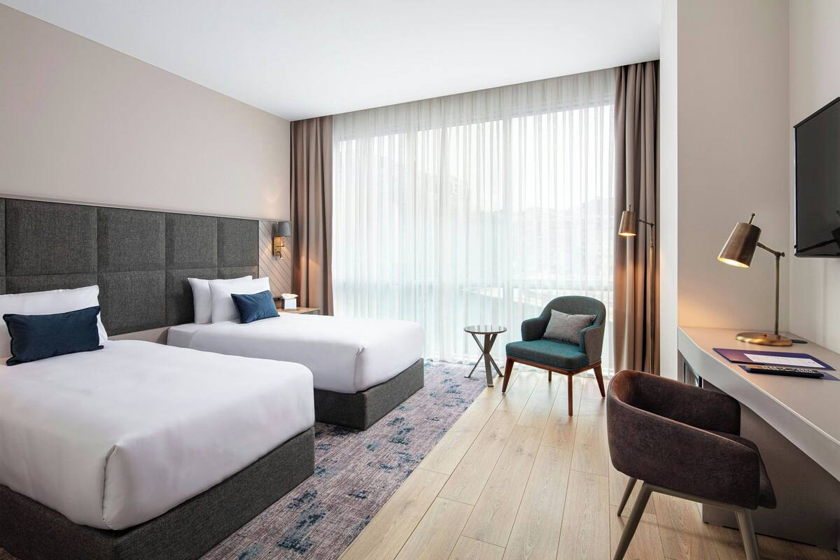 A hotel room with two beds, a desk, a chair, a TV, and a large window with curtains.