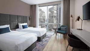 Quarto de hotel com duas camas, uma escrivaninha, uma cadeira e uma janela grande com vista para a cidade.