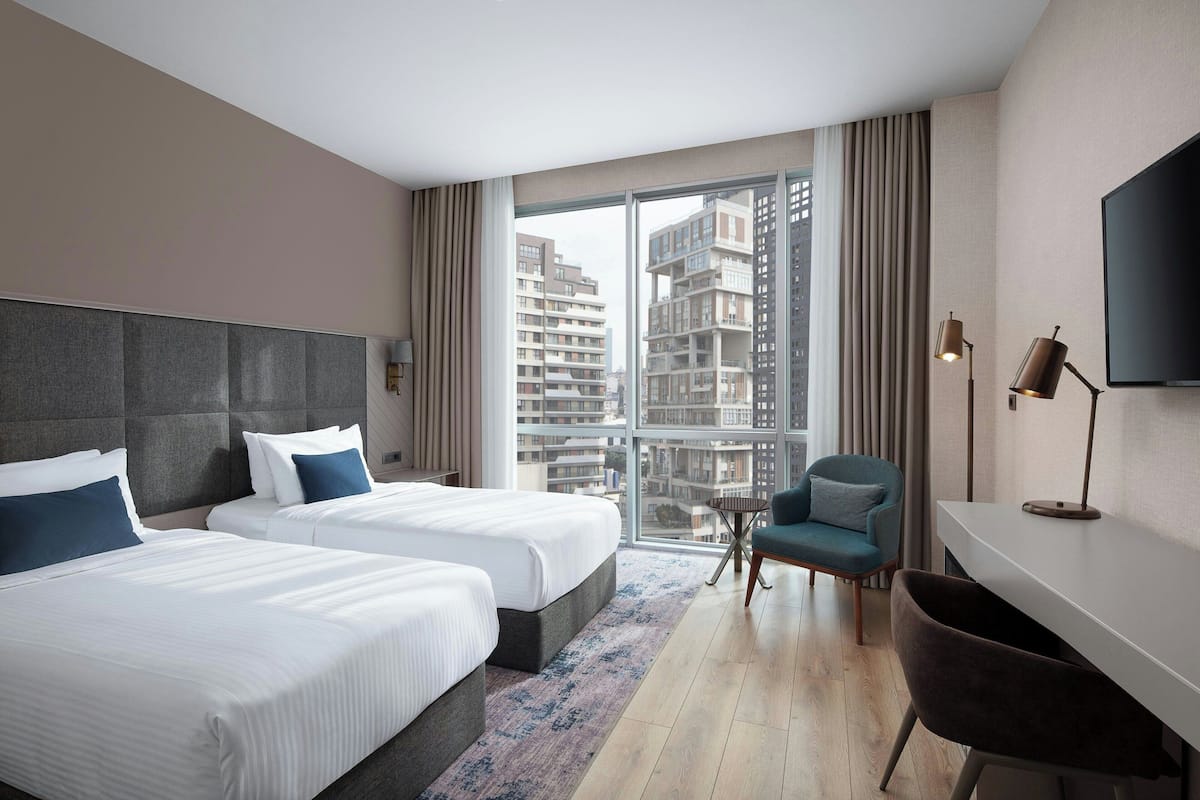 A hotel room with two beds, a desk, a chair, and a large window offering a city view.