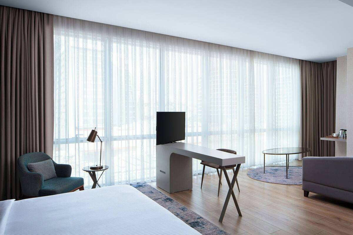 A hotel room with a bed, a desk with a TV, a chair, a sofa, and large windows with curtains.
