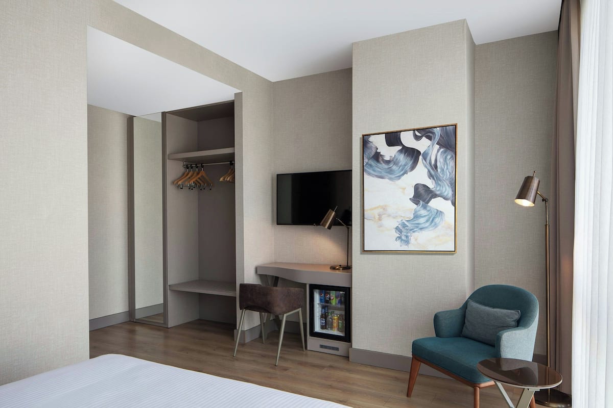 A modern hotel room with a bed, a desk with a TV, a chair, a coat rack, and a painting on the wall.