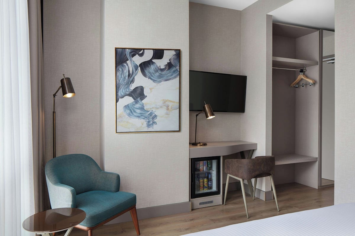 A modern hotel room with a teal armchair, a small table, a flat-screen TV, and a built-in mini-fridge.