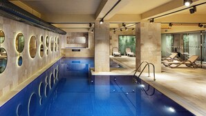 Uma piscina interior moderna com piso em azulejos azuis, cadeiras de descanso e uma parede com espelhos circulares.