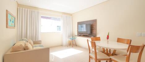 A modern living room with a sofa, a dining table with chairs, a TV mounted on the wall, and a window with curtains.