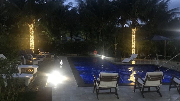 Un espace aménagé au bord de la piscine, avec des chaises longues et une piscine, le tout entouré de palmiers et illuminé par des guirlandes lumineuses.
