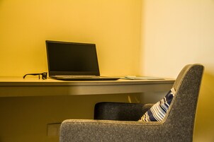 Een laptop op een bureau met een paar brillen ernaast, een stoel met een gestreept kussen, en een effen muur op de achtergrond.