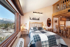A bedroom with a large bed, a wooden headboard, a view of mountains, a dining area with wooden furniture, and a kitchen with wooden cabinets. - Log Cabin in the Lodge | AC, Pool, Hot Tub, Golf & Ski (Durango)