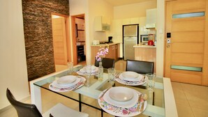 A dining area with a glass table set for four, surrounded by chairs. A kitchen is visible in the background with modern appliances. - Praia Apartments by Casago (Playa del Carmen)