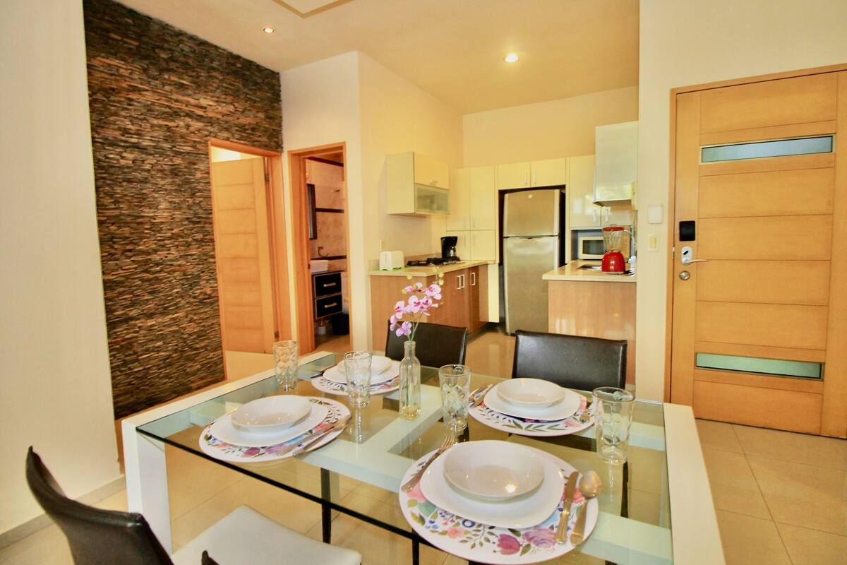 A dining area with a glass table set for four, surrounded by chairs. A kitchen is visible in the background with modern appliances.
