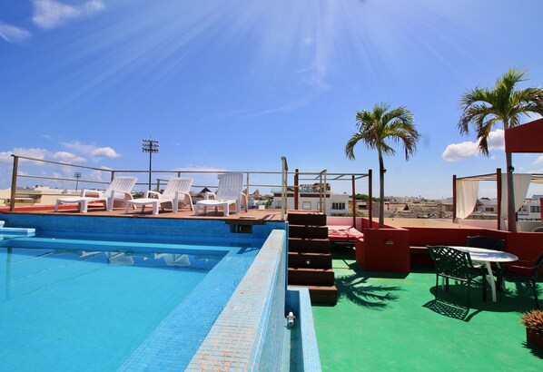 Uma piscina no terraço com cadeiras de descanso brancas, uma mesa e vista para a cidade.