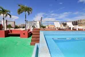 Uma área de piscina no terraço com espreguiçadeiras brancas, palmeiras e um céu azul límpido.
