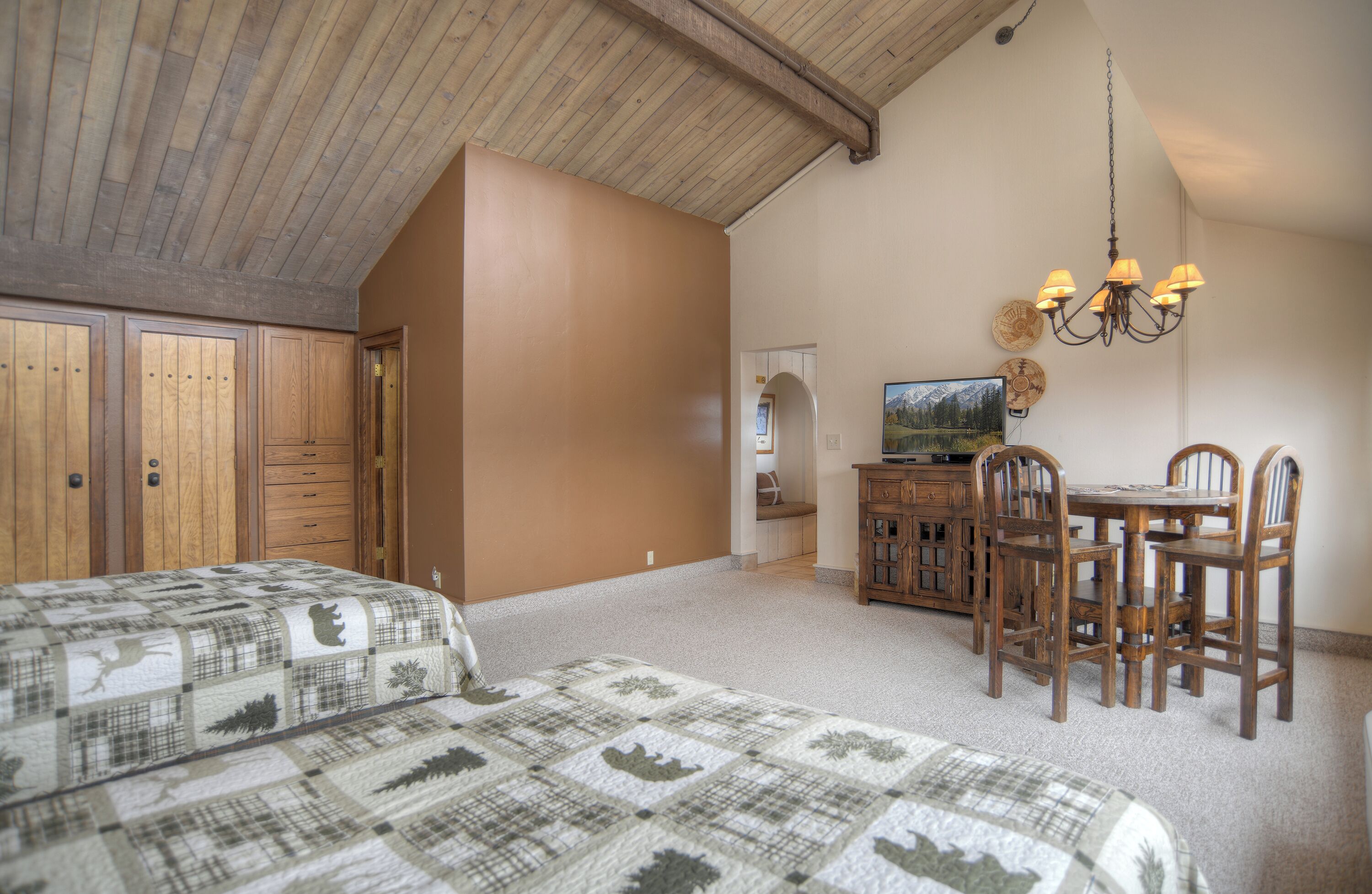 A bedroom with two beds, a wooden ceiling, a dining table with chairs, and a chandelier.