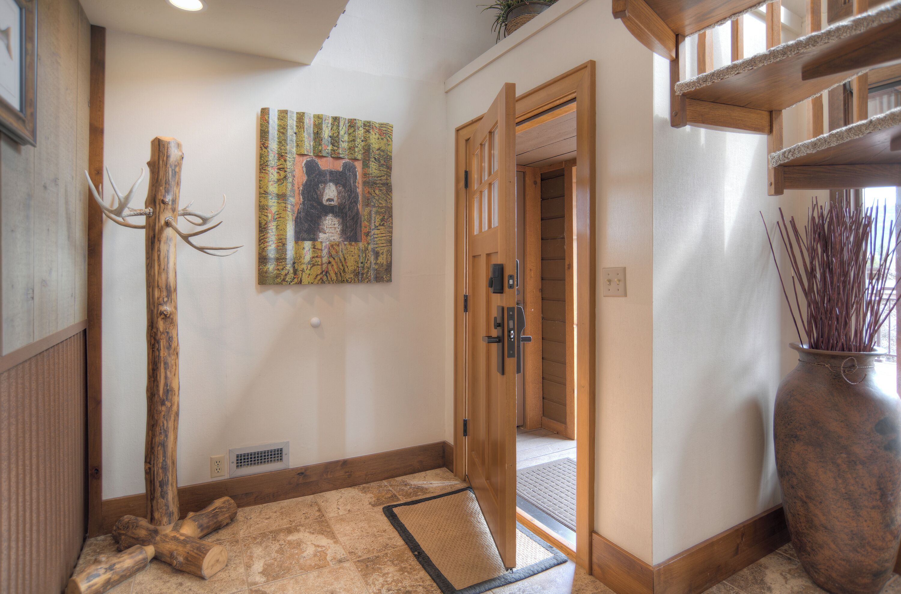 A wooden door with a glass panel, a bear painting on the wall, and a rustic decor with a wooden post and a vase of reeds.