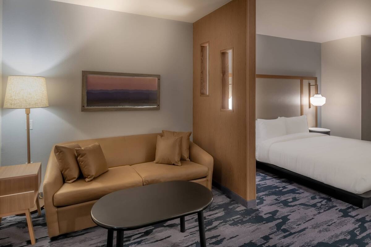 Une chambre d’hôtel avec un canapé, une petite table, un lit, une table de chevet, une lampe et un tableau au mur.