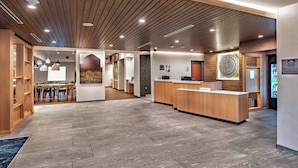 Un hall d’hôtel moderne avec un comptoir d’accueil en bois, un espace salon avec des tables et une grande œuvre d’art murale.