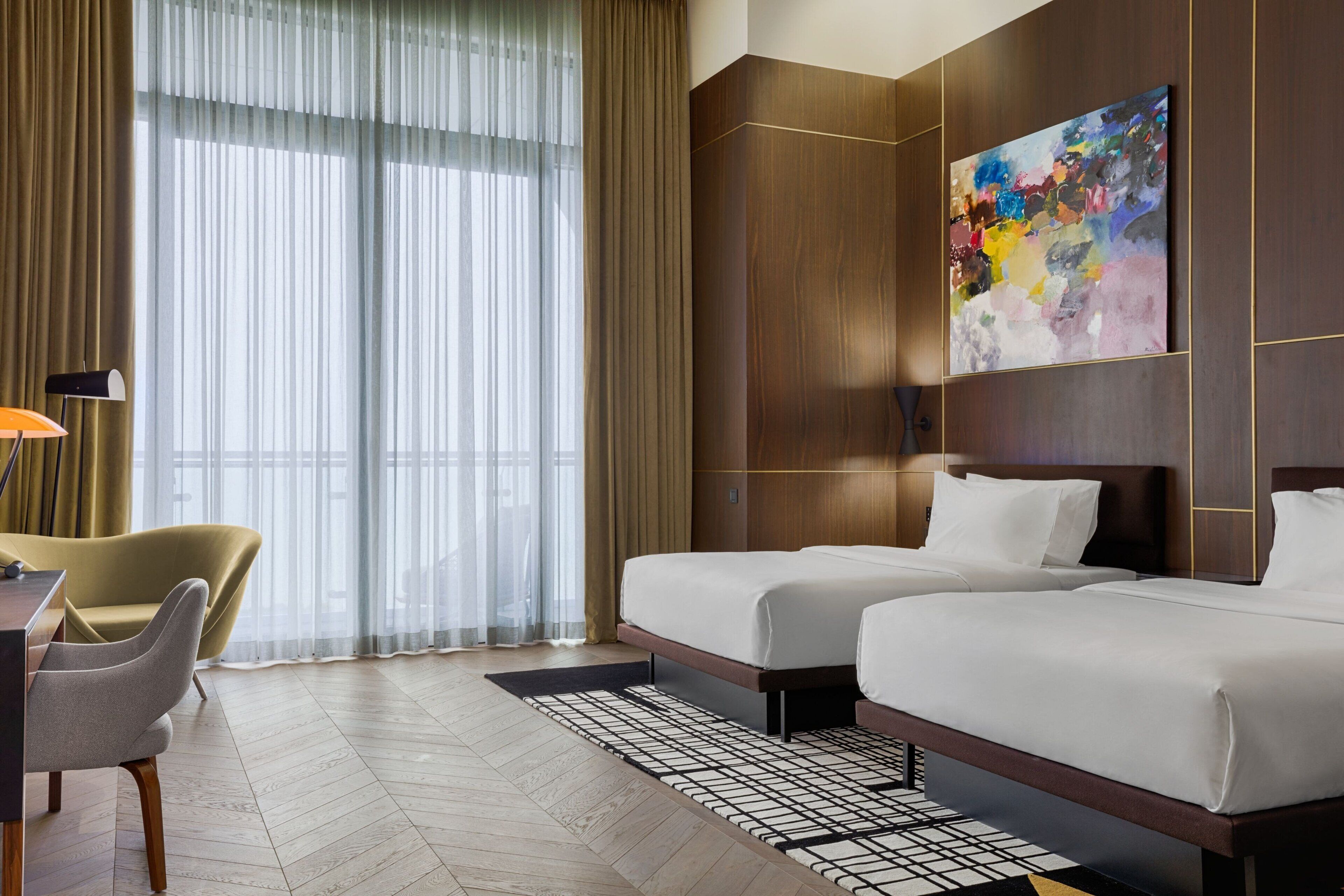Een hotelkamer met twee bedden, een bureau, een stoel, een groot raam met dunne gordijnen en een kleurrijk abstract schilderij aan de muur.