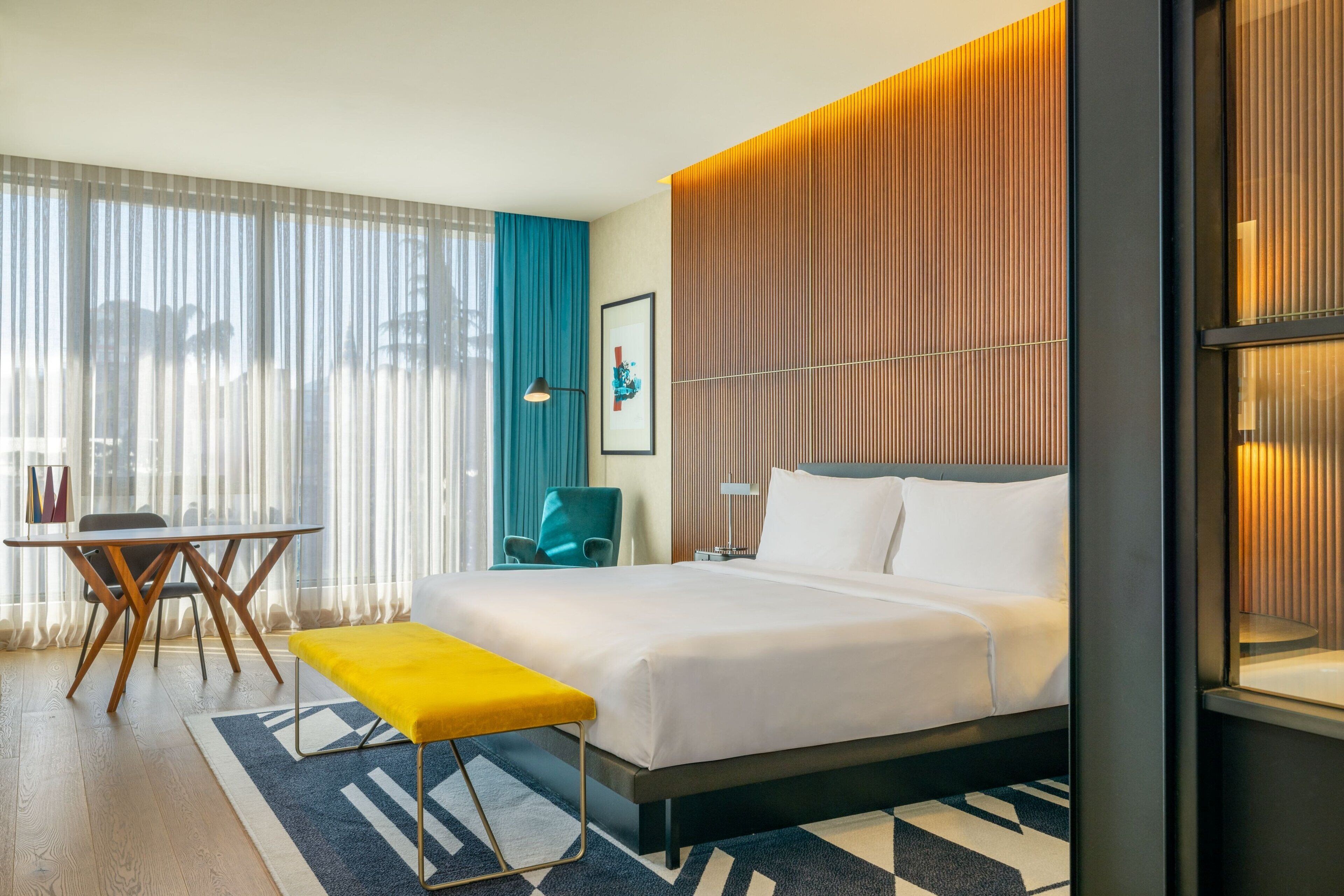A modern hotel room with a large bed, a desk with chairs, a bench, and a view of the outdoors through large windows.