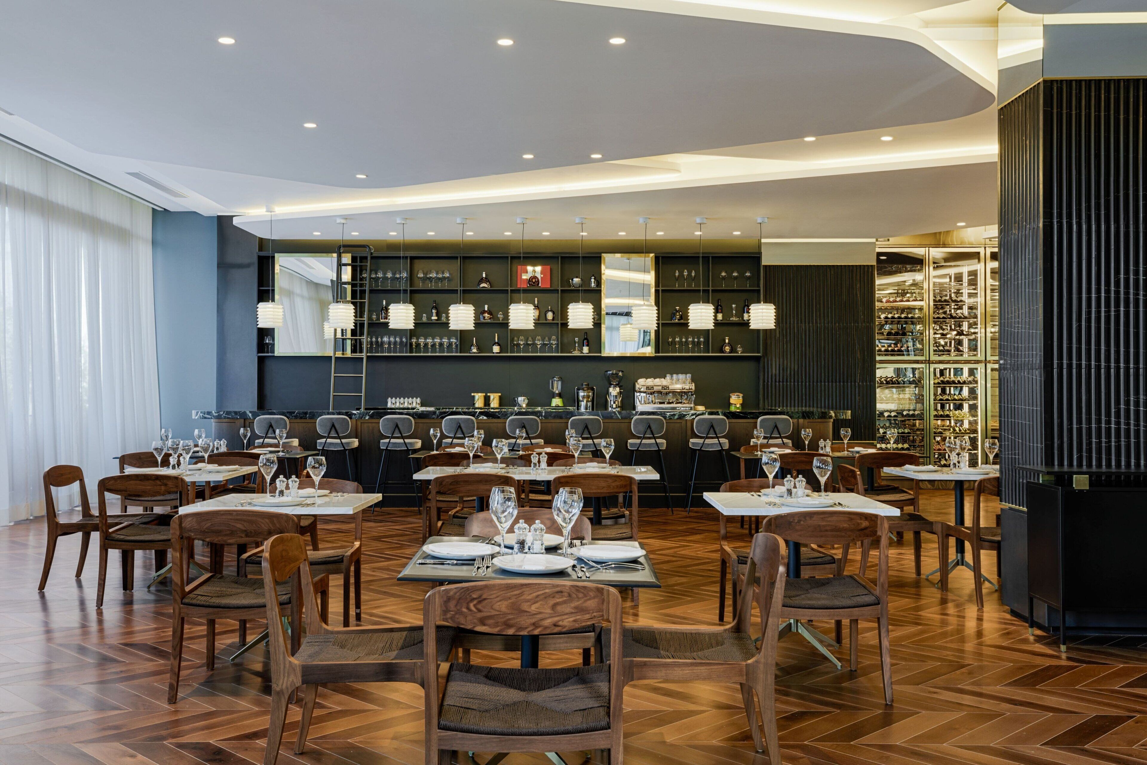 A restaurant with wooden flooring, tables set with white tablecloths, and a bar area with shelves of bottles.