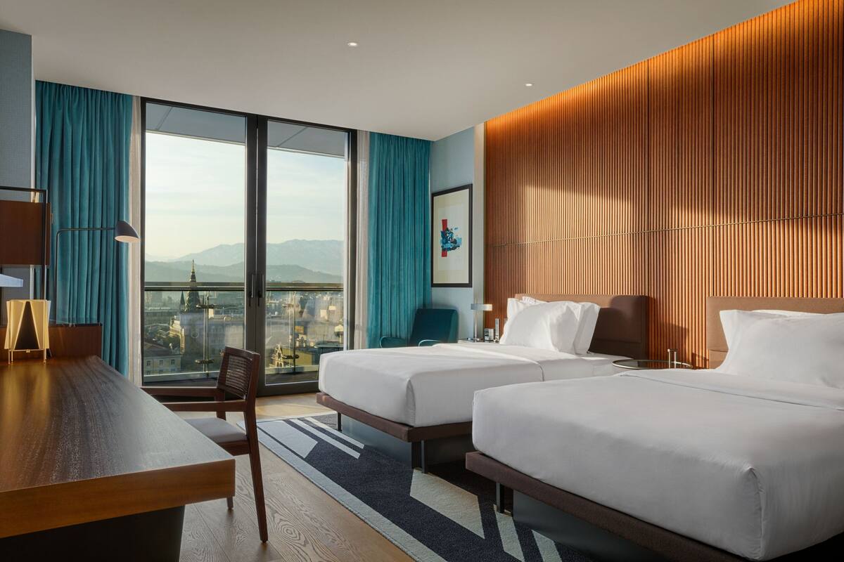 A hotel room with two beds, a desk, a chair, a large window with a city view, and a wooden wall panel.