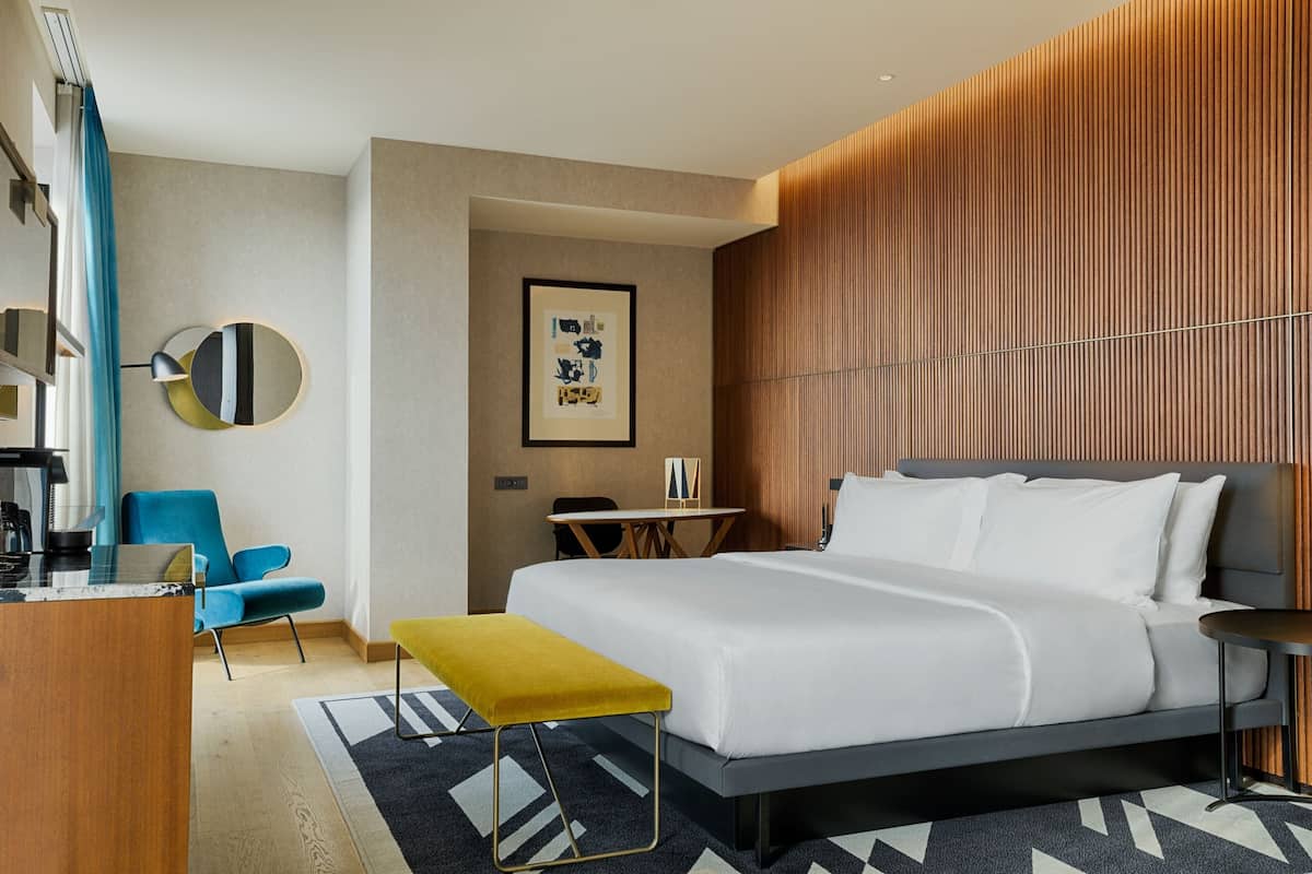 A modern hotel room with a large bed, a desk, a chair, and a small table.