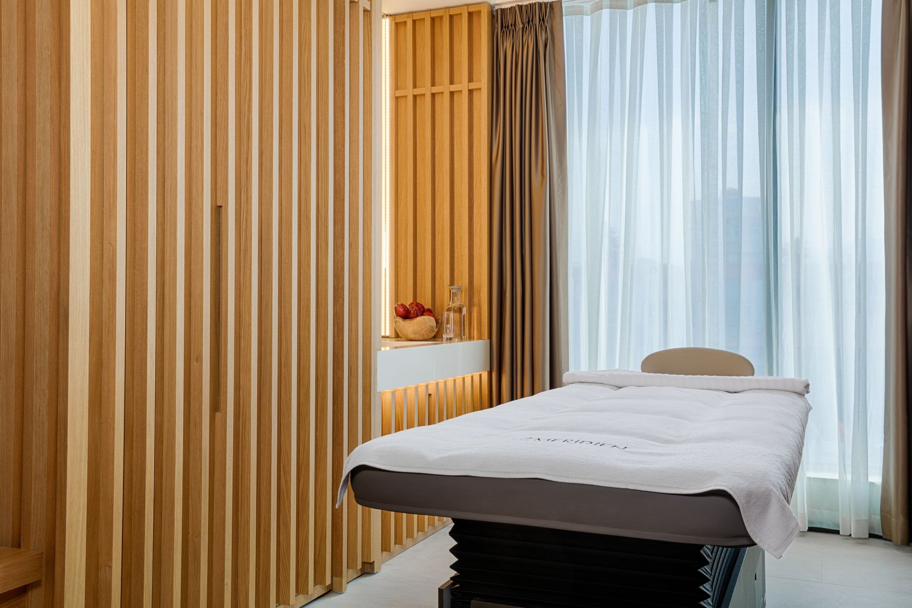 A modern hotel room with a wooden slatted wall, a bed with a white mattress, and a view through sheer curtains.