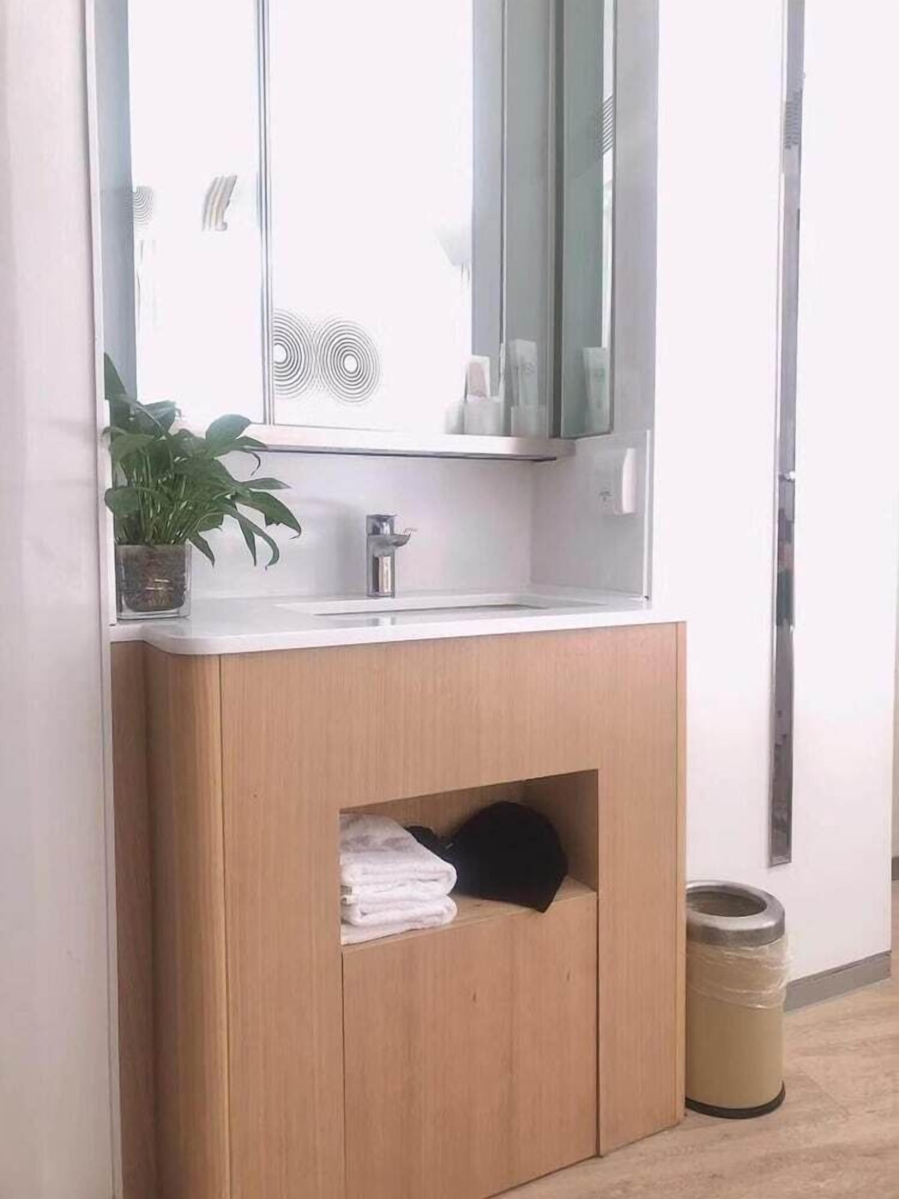 A bathroom with a wooden vanity, a sink, a mirror, a potted plant, and a trash can.