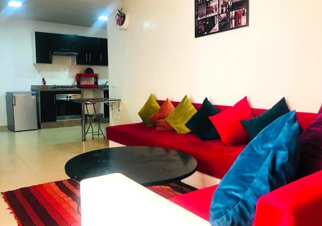 A living room with a red sofa, black coffee table, and colorful cushions. There is a kitchen in the background with black cabinets and a red appliance.