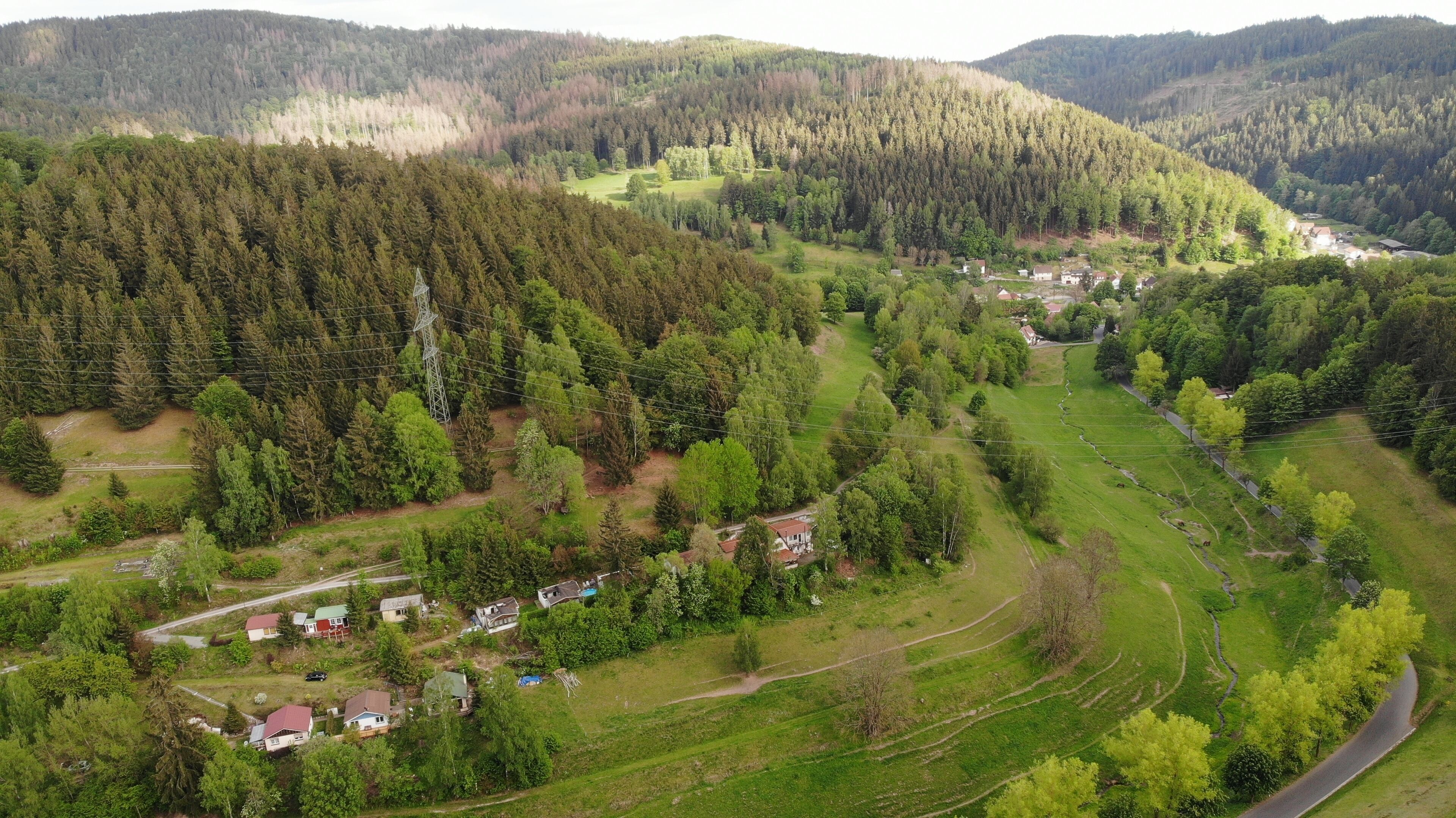 Eine Vogelperspektive auf eine ländliche Gegend mit verstreuten Häusern, einer kurvenreichen Straße und dicht bewaldeten Hügeln im Hintergrund.