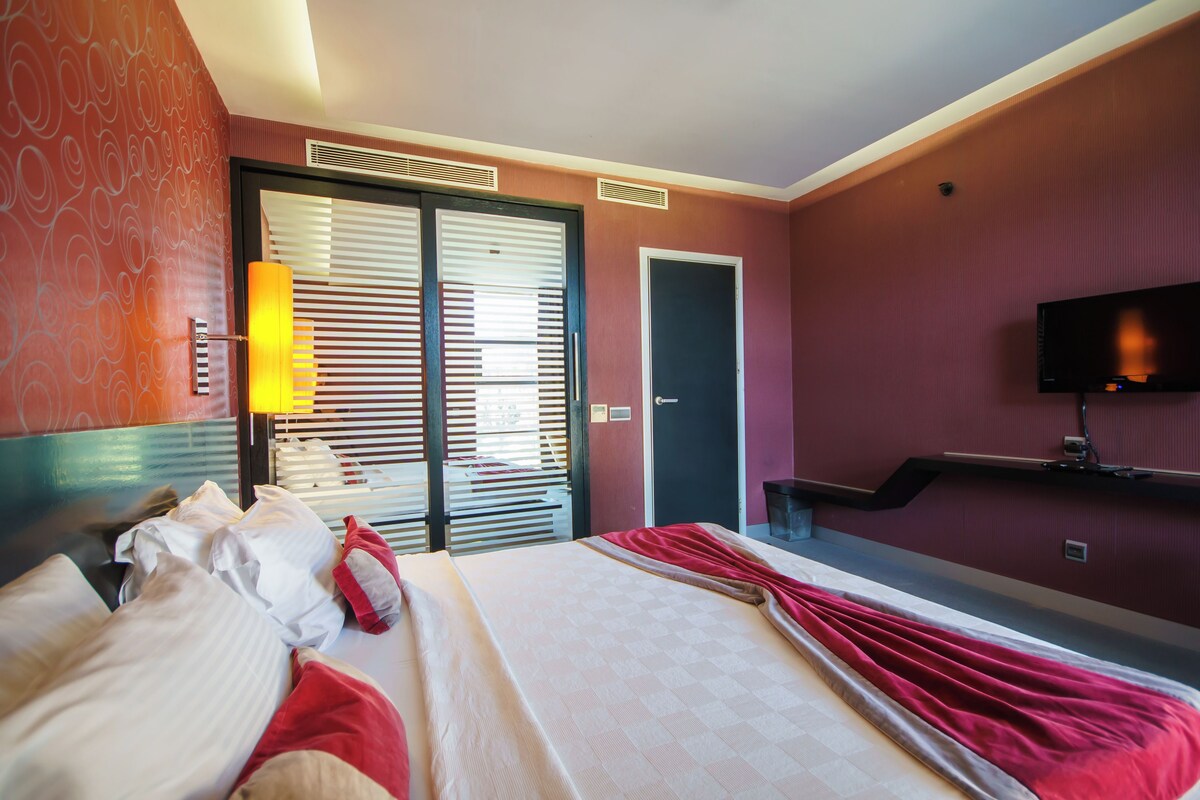 A hotel room with a bed, a television, a door, and a window with blinds.