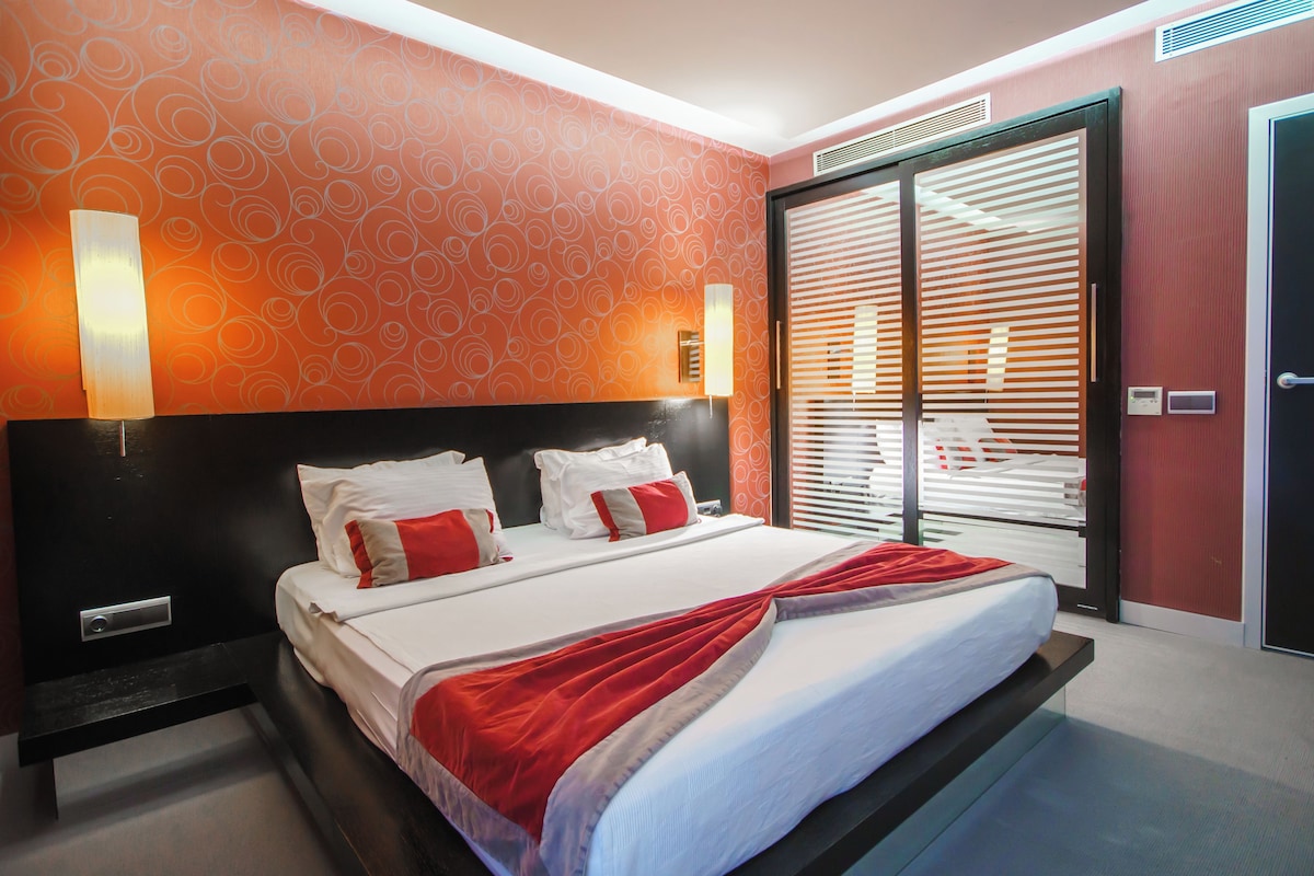 Una habitación de hotel con cama, mesita de noche, lámpara de pared, puerta con manija y ventana con persianas.