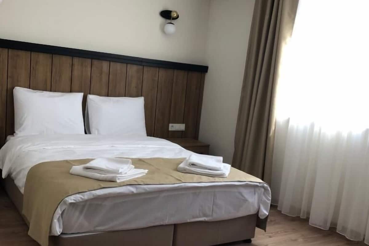 A neatly made bed with white linens and a beige blanket, a wooden headboard, a bedside table with a lamp, and a large window with curtains.