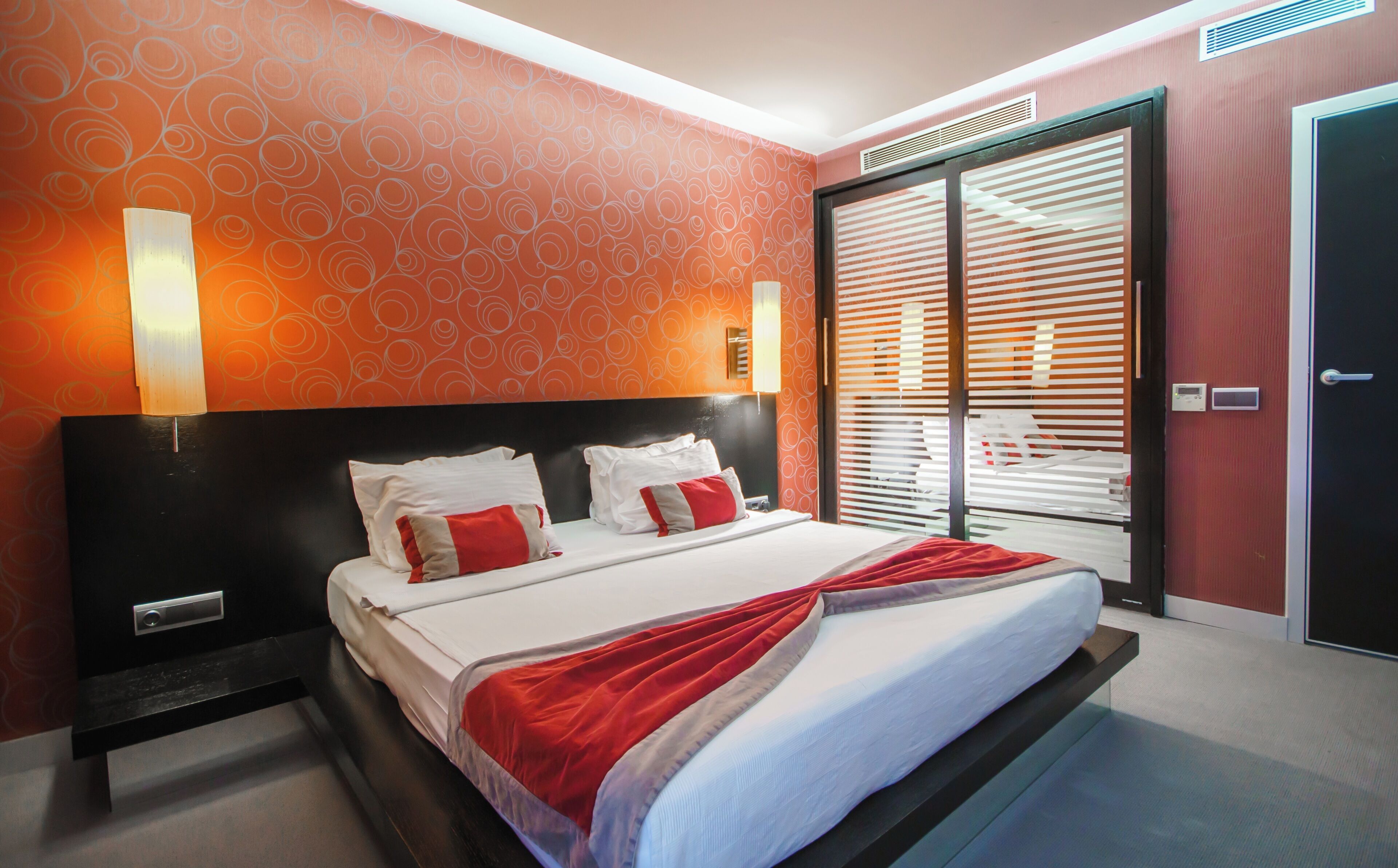 A hotel room with a bed, a nightstand, a wall-mounted lamp, a door with a handle, and a window with blinds.