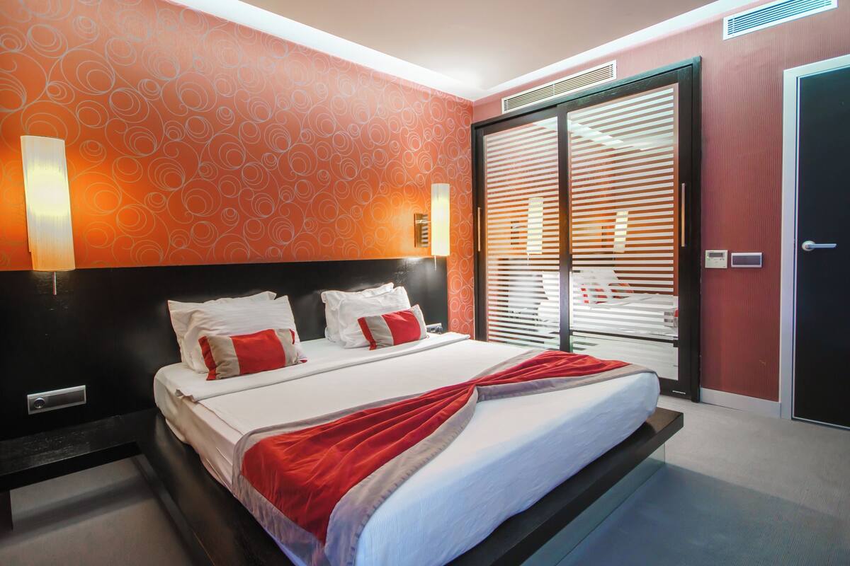 A hotel room with a bed, a nightstand, a wall-mounted lamp, a door with a handle, and a window with blinds.