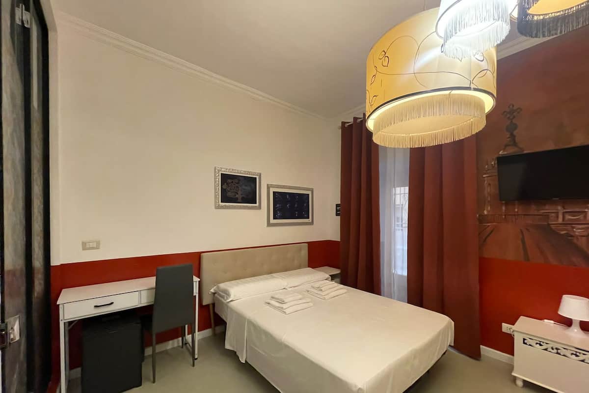 A hotel room with a bed, a desk, a chair, a TV, and a chandelier.
